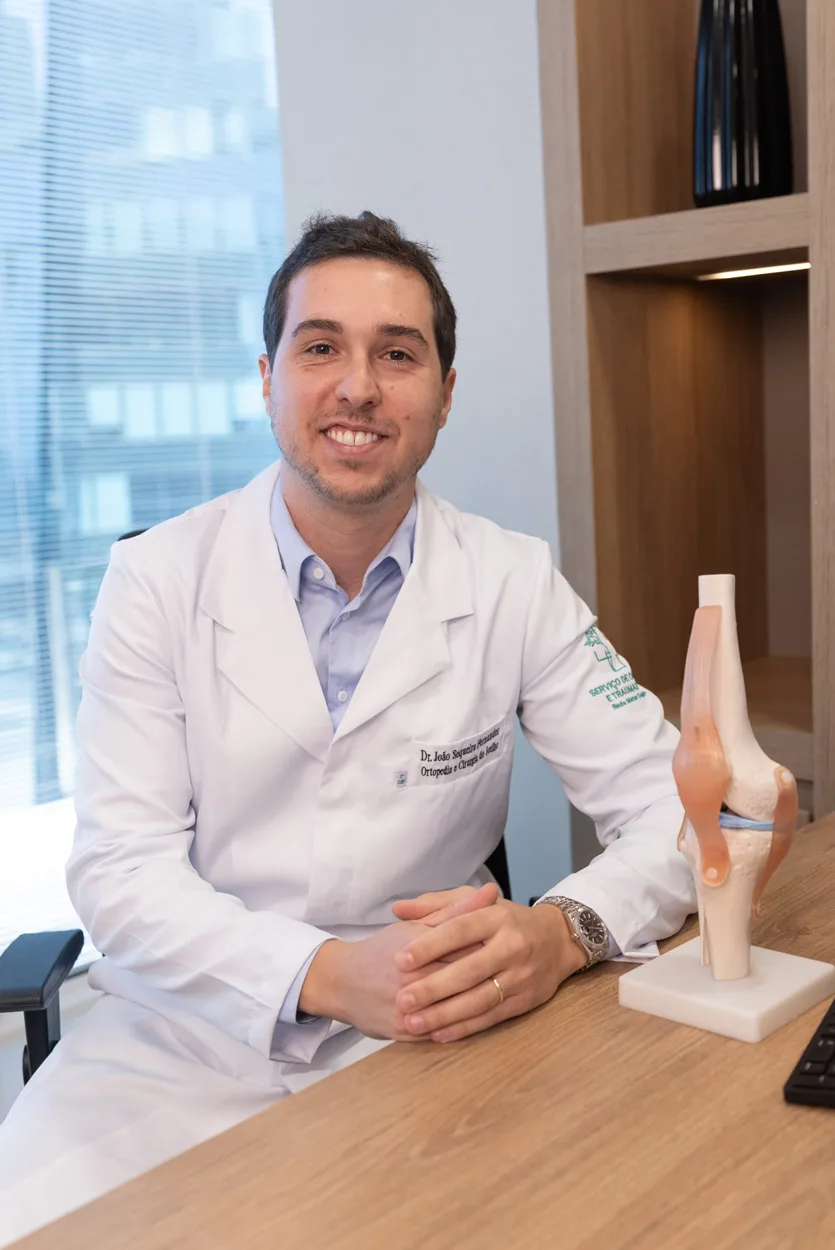 Dr. João Sequeira, ortopedista em Belo Horizonte, sentado à mesa ao lado de um modelo anatômico do joelho